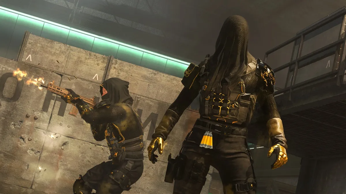 An operator in black and gold with a black hood stands in front of another operator shooting.
