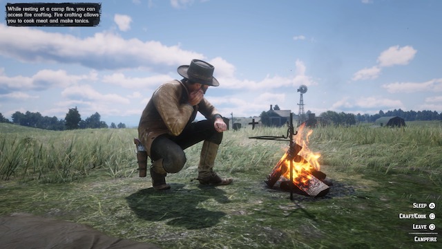 Arthur kneels next to a campfire in a field near a farmhouse in RDR2